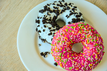 An appetizing donut with pink icing and a multi-colored sprinkle and a delicious chocolate donut with white icing sprinkled with chocolate chips and on a white plate