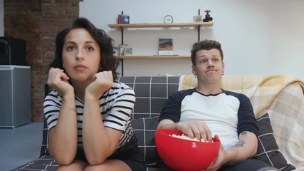Unhappy gorgeous Caucasian couple in love sitting on couch and watching at broken TV. Guy throwing popcorn. Unlucky day. Indoors.