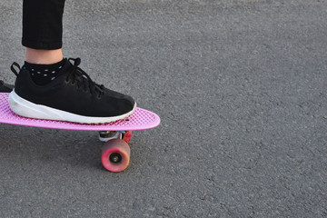 One foot in black sneakers on pink skateboard or penny board on gray asphalt background with copy space for text.