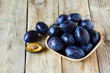 Ripe blue plums with water drops in a wooden bowl on a wooden background. Half a plum with a stone