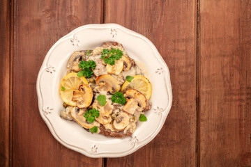 A photo of scallopine di vitello, veal scallopini, with a mushroom sauce, lemons, and parsley, shot from above on a dark rustic wooden table with a place for text