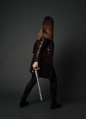 full length portrait of brunette girl wearing long leather coat and boots. standing pose and holding a sword on grey studio background.