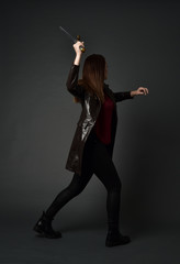 full length portrait of brunette girl wearing long leather coat and boots. standing pose and holding a sword on grey studio background.