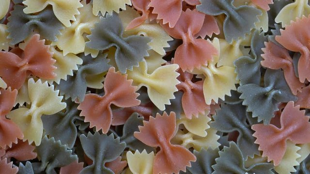 Multicolored pasta background, rotating, top view. Rotation of colored farfalle pasta. Heap of uncooked pasta. Italian food close up. Delicious italian farfalle tricolor pasta, food background