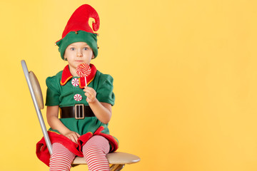 A beautiful baby in a suit of a Christmas elf is sitting on a chair. She has a delicious lollipop in her hands. Surprise. Near Copy Space.