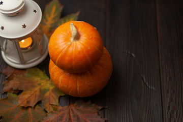 Two orange pumpkins on a dark brown wooden background. Is a decorative lantern with a burning candle. Near lie maple leaves. Autumn composition. Halloween. Harvest. Postcard
