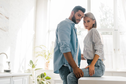 side view of passionate young couple hugging in kitchen, girlfriend looking at camera
