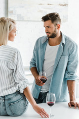 Obraz premium young couple looking at each other, boyfriend holding glass of wine in kitchen