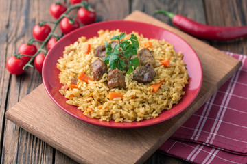 Red plate with pilaf on a brown wooden table. On the table is red pepper, green onions, garlic, cherry tomatoes, red napkin, spoon.