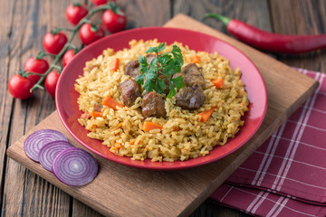 Red plate with pilaf on a brown wooden table. On the table is red pepper, green onions, garlic, cherry tomatoes, red napkin, spoon.