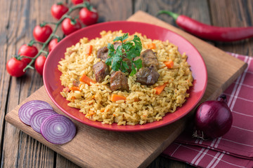 Red plate with pilaf on a brown wooden table. On the table is red pepper, green onions, garlic, cherry tomatoes, red napkin, spoon.
