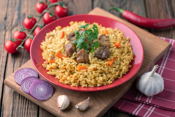 Red plate with pilaf on a brown wooden table. On the table is red pepper, green onions, garlic, cherry tomatoes, red napkin, spoon.