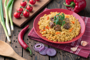 Red plate with pilaf on a brown wooden table. On the table is red pepper, green onions, garlic, cherry tomatoes, red napkin, spoon.