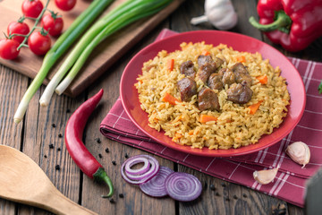 Red plate with pilaf on a brown wooden table. On the table is red pepper, green onions, garlic, cherry tomatoes, red napkin, spoon.
