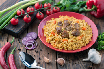 Red plate with pilaf on a brown wooden table. On the table is red pepper, green onions, garlic, cherry tomatoes, red napkin, spoon.