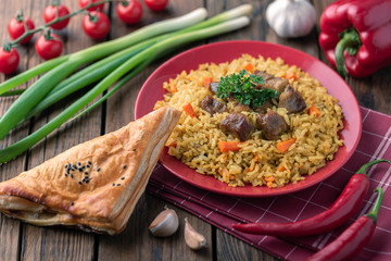 Red plate with pilaf on a brown wooden table. On the table is red pepper, green onions, garlic, cherry tomatoes, red napkin, spoon.