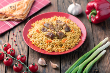 Red plate with pilaf on a brown wooden table. On the table is red pepper, green onions, garlic, cherry tomatoes, red napkin, spoon.