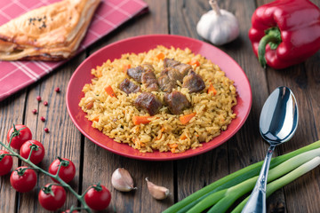 Red plate with pilaf on a brown wooden table. On the table is red pepper, green onions, garlic, cherry tomatoes, red napkin, spoon.