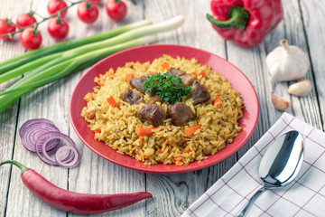 Red plate with pilaf on a white wooden table. On the table is red pepper, green onions, garlic, cherry tomatoes, white napkin, spoon.
