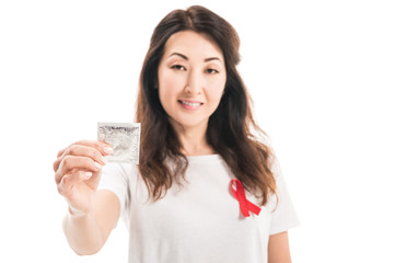 smiling adult asian woman with aids awareness red ribbon on t-shirt holding condom isolated on white