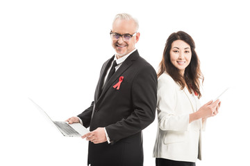 Fototapeta premium multiethnic happy businesspeople with aids awareness red ribbons working with gadgets while standing back to back and looking at camera isolated on white