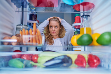 Confused woman choosing what to eat. Hands on head. Picture taken from the inside of fridge full of...