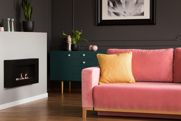 Yellow pillow on a powder pink, velvet sofa and a black, eco burning fireplace in a modern vintage living room interior with dark walls. Real photo.