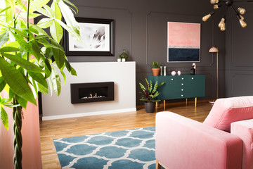 Green plants in a hipster living room interior with molding on dark walls and a pink sofa in front of a burning fireplace. Real photo.