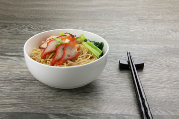 Egg noodle with red roast pork and vegetables in white bowl and black chopstick on wood table background..
