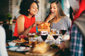 Friends giving gifts to each other while sitting at table. In background Christmas tree. Christmas holidays concept.