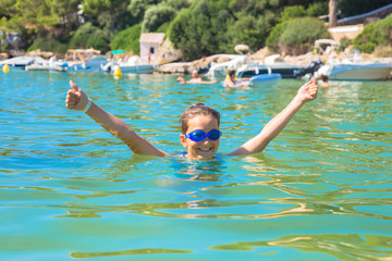 Happy child girl swims in the sea and shows a thumbs up symbol.