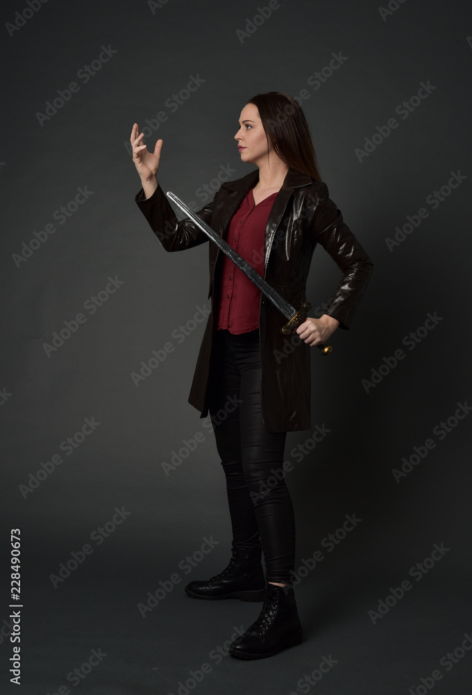 Poster full length portrait of brunette girl wearing long leather coat and boots. standing pose and holding a sword on grey studio background.