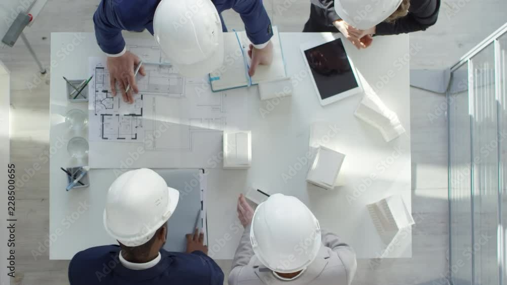 Poster top view of four multiethnic engineers in protective helmets using scale models and blueprints while