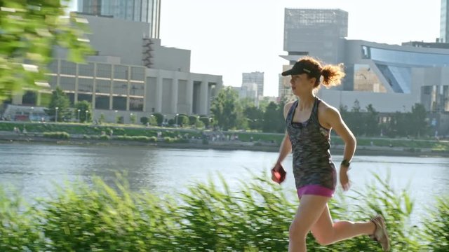 Tracking shot of muscly female athlete wearing cap jogging along embankment on sunny summer day