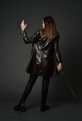 full length portrait of brunette girl wearing long leather coat and boots. standing pose and holding a sword on grey studio background.