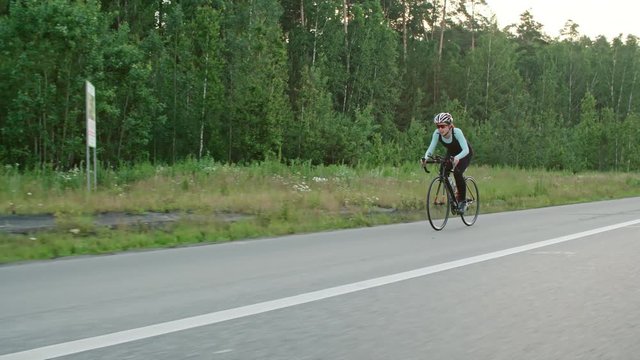 Tracking of female cyclist in protective helmet and professional sportswear riding bicycle along road surrounded by green forest