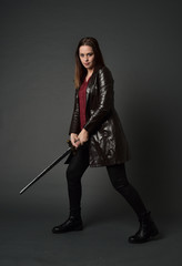 full length portrait of brunette girl wearing long leather coat and boots. standing pose and holding a sword on grey studio background.