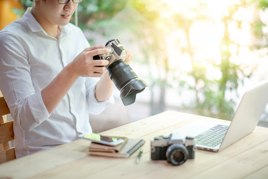 Young Asian man holding digital single-lens reflex camera (DSLR) checking photo on camera screen display sitting with laptop computer and smartphone on the table. Photography, art and hobby concepts