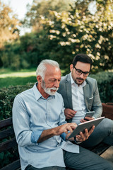 Fototapeta premium Senior businessman giving advice to a younger colleague outdoors. Looking at tablet while sitting on the bench.