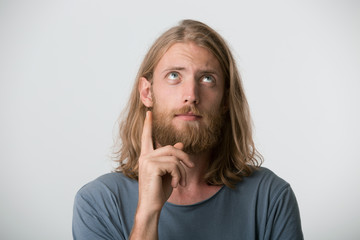 Young guy with a beard, mustache, blond hair to the shoulders and blue eyes looks up feels contemplative, focused and having some idea. Points up with one finger, isolated over white background