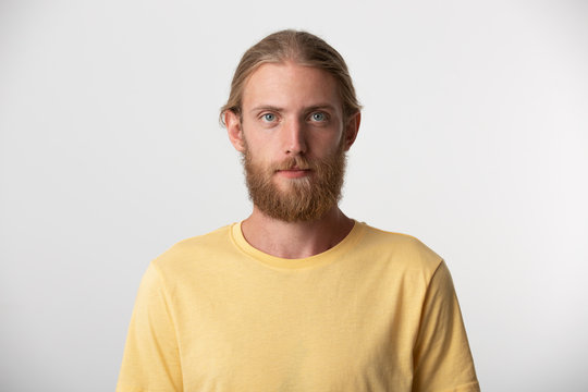 A young guy with a beard, mustache, blond hair put back and blue eyes feels calm confident looking camera with day to day expression, wears yellow tshirt isolated over white background