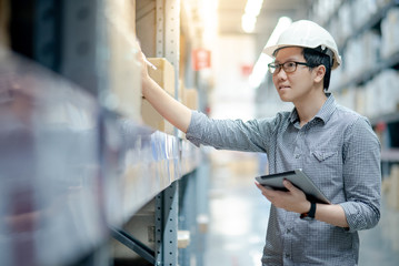 Young Asian man worker wearing safety helmet and eyeglasses doing stocktaking of product in cardboard box on shelves in warehouse by using digital tablet and pen. Physical inventory count concept