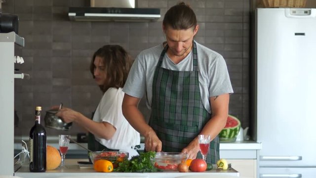 Husband and wife in the kitchen preparing dinner. He cuts the salad, she cooks the soup on the stove in the pan. Girl brings the pan, removes the lid, guy throws a piece of pepper there. Family humor