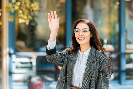 smiling woman in stylish jacket waving to someone on street