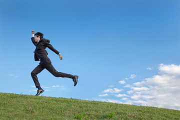 青空をバックに坂道を駆け上るスーツ姿の若いビジネスマン1人の横姿。競争・目標・元気・挑戦イメージ