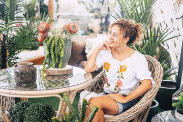 Obraz premium Young woman with blond curly hair sitting on a wicker chair with many plants admires the vegetation. Relaxation and leisure concept. green colors and people