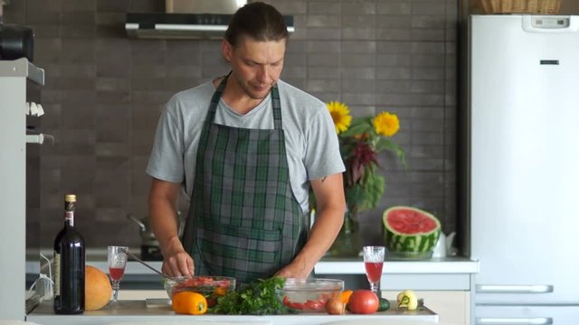 A man in an apron in the kitchen. The guy is fooling around with a serious face. Throws pepper like a grenade. Hiding under the table