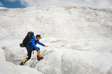 A mountaineer with a backpack walks in crampons walking along a dusty glacier with sidewalks in the hands between cracks in the mountain