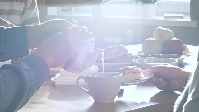 Mid-section shot with PAN of unrecognizable young man pouring hot tea into cup of senior lady, then taking piece of pie from woman while having breakfast