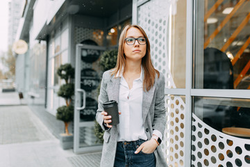 Girl blogger with coffee standing on the street, waiting for a meeting. Stylish and smiling girl. A happy buyer, a modern coffee shop.
